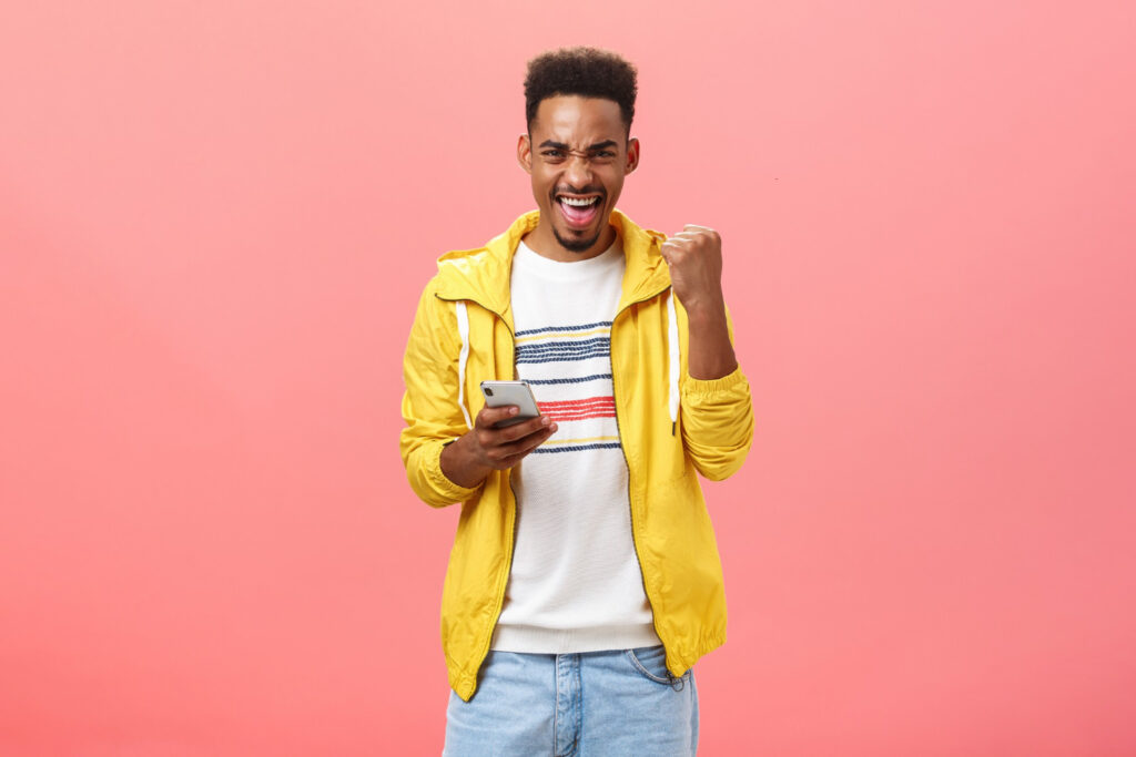 Portrait of happy male raising fist in victory and triumph gesture winning in online phone game over pink wall.